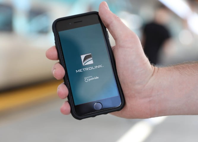 Phone being held in a hand with the Metrolink app start screen on it in the background of the scene is an out of focus train station